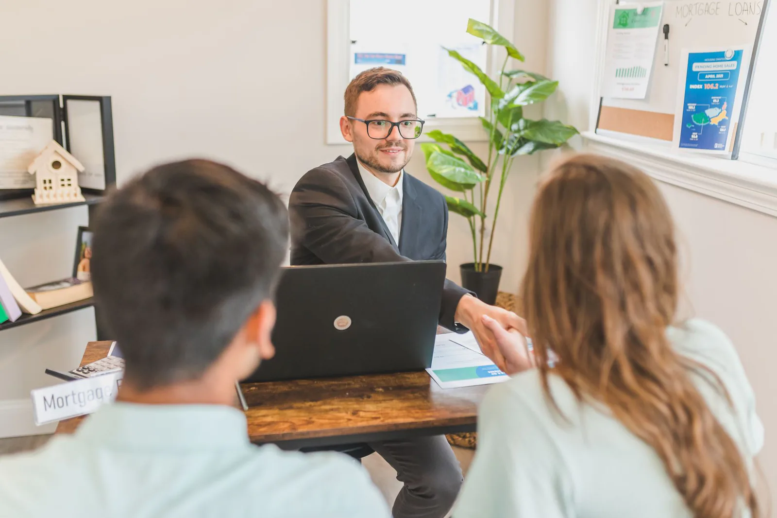 Mortgage broker shaking hands with a couple of buyers in his office, in front of a laptop